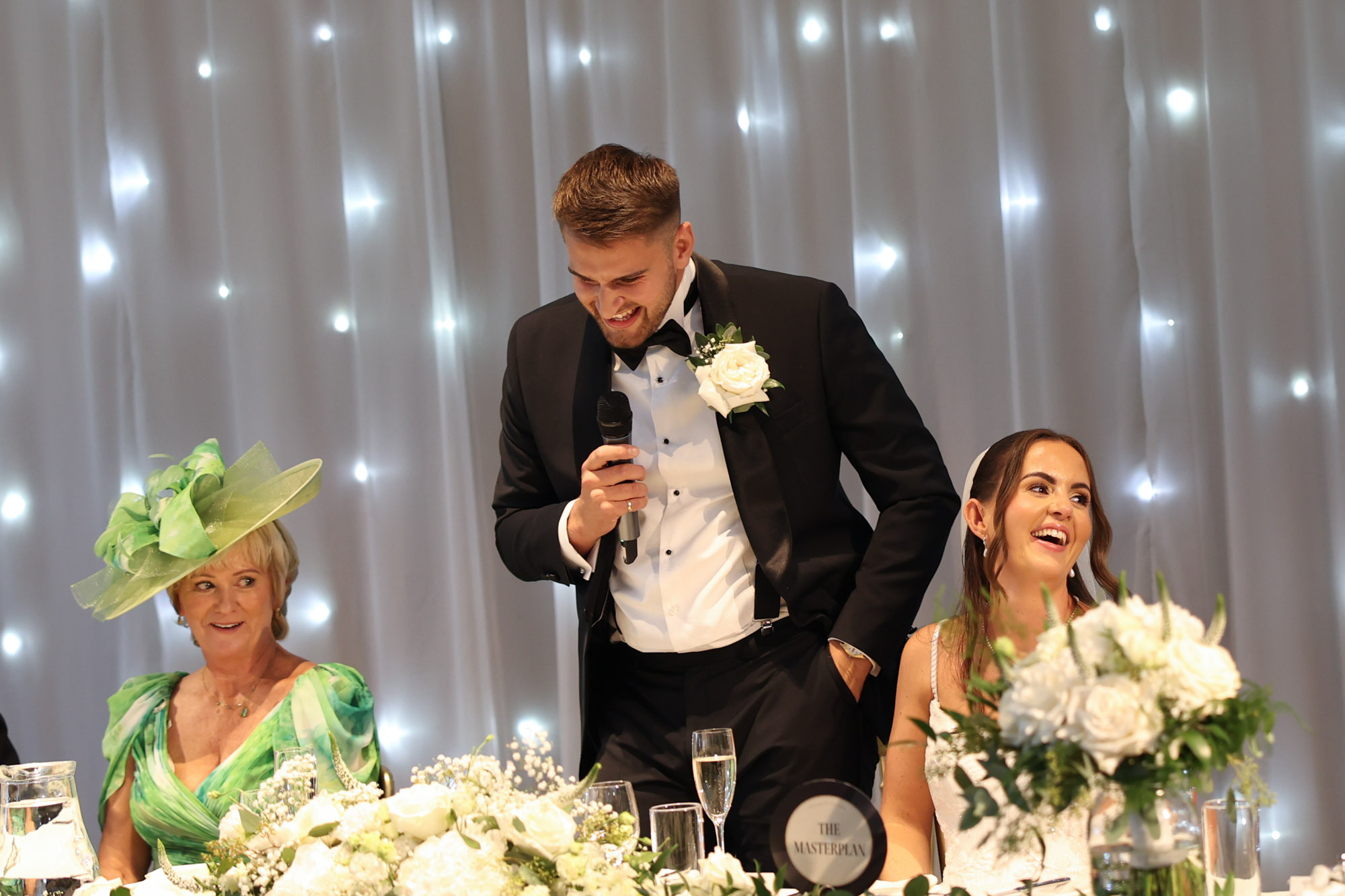 A groom gives a speech at the top table beside a laughing bride and a guest wearing a green dress.