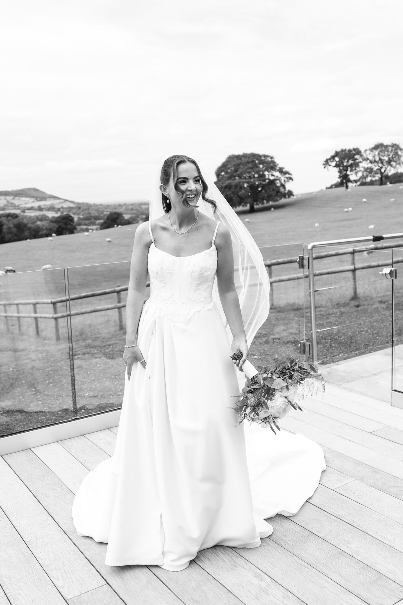 Black and white photo of a bride in a gown and veil holding a bouquet on a wooden deck with countryside views.