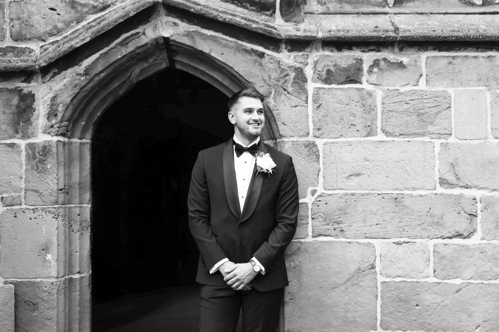 Black and white portrait of a smiling groom in a tuxedo standing in front of an arched stone doorway.