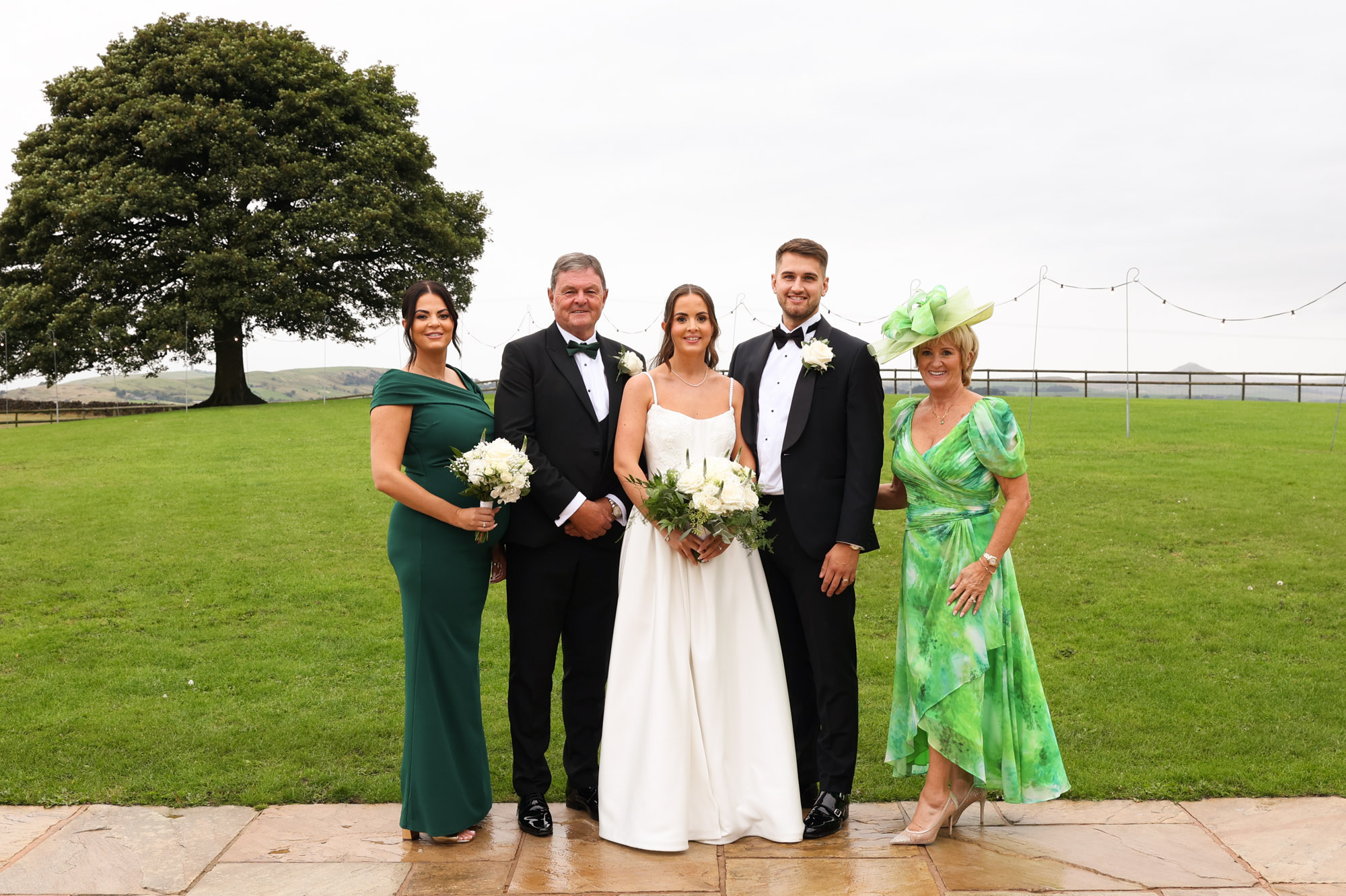 Bride and groom pose outdoors with three family members in green attire and tuxedos, with an oak tree in the background.