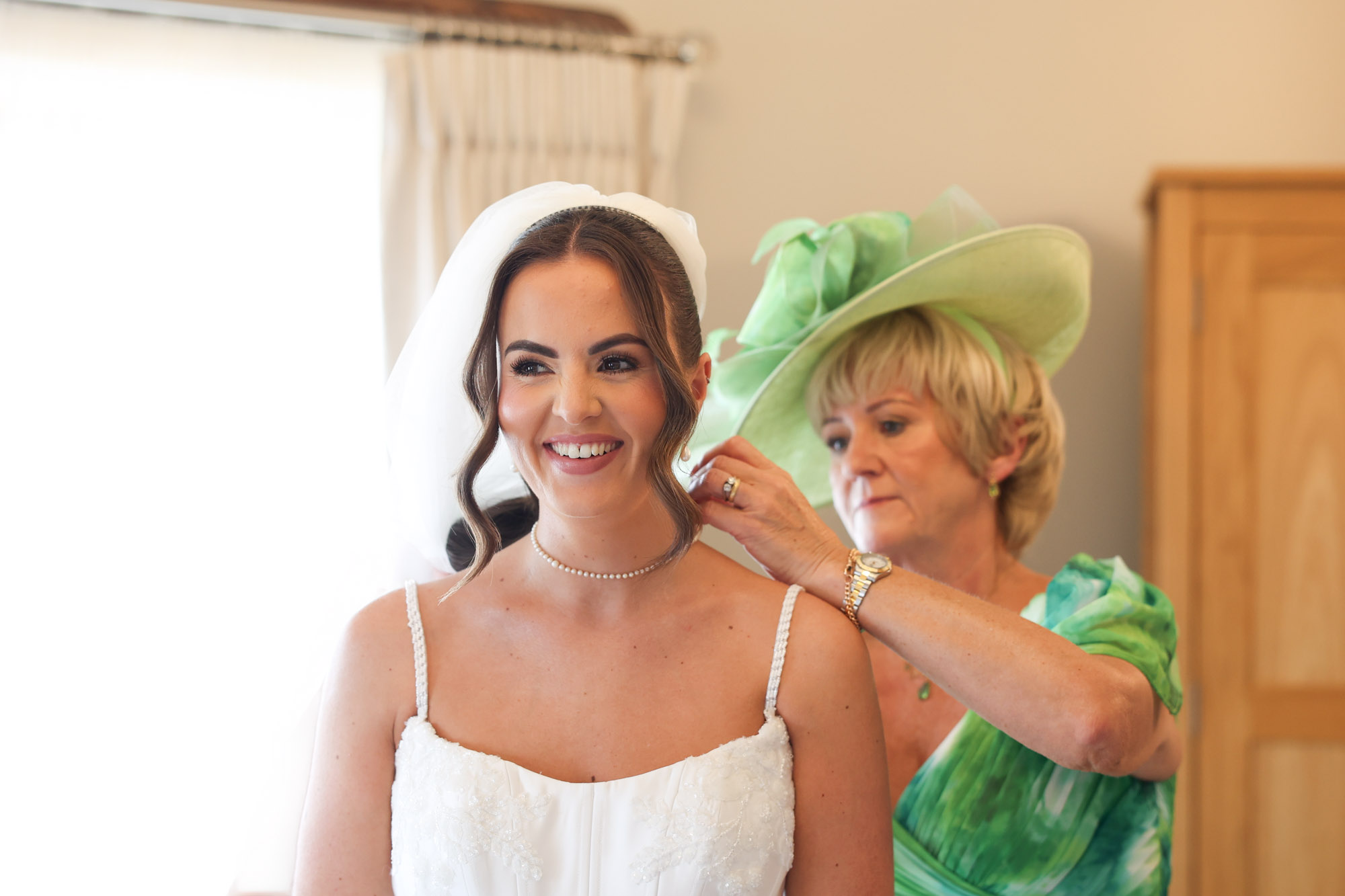 A mother in a green dress and matching hat adjusts the veil on the smiling bride during prep.
